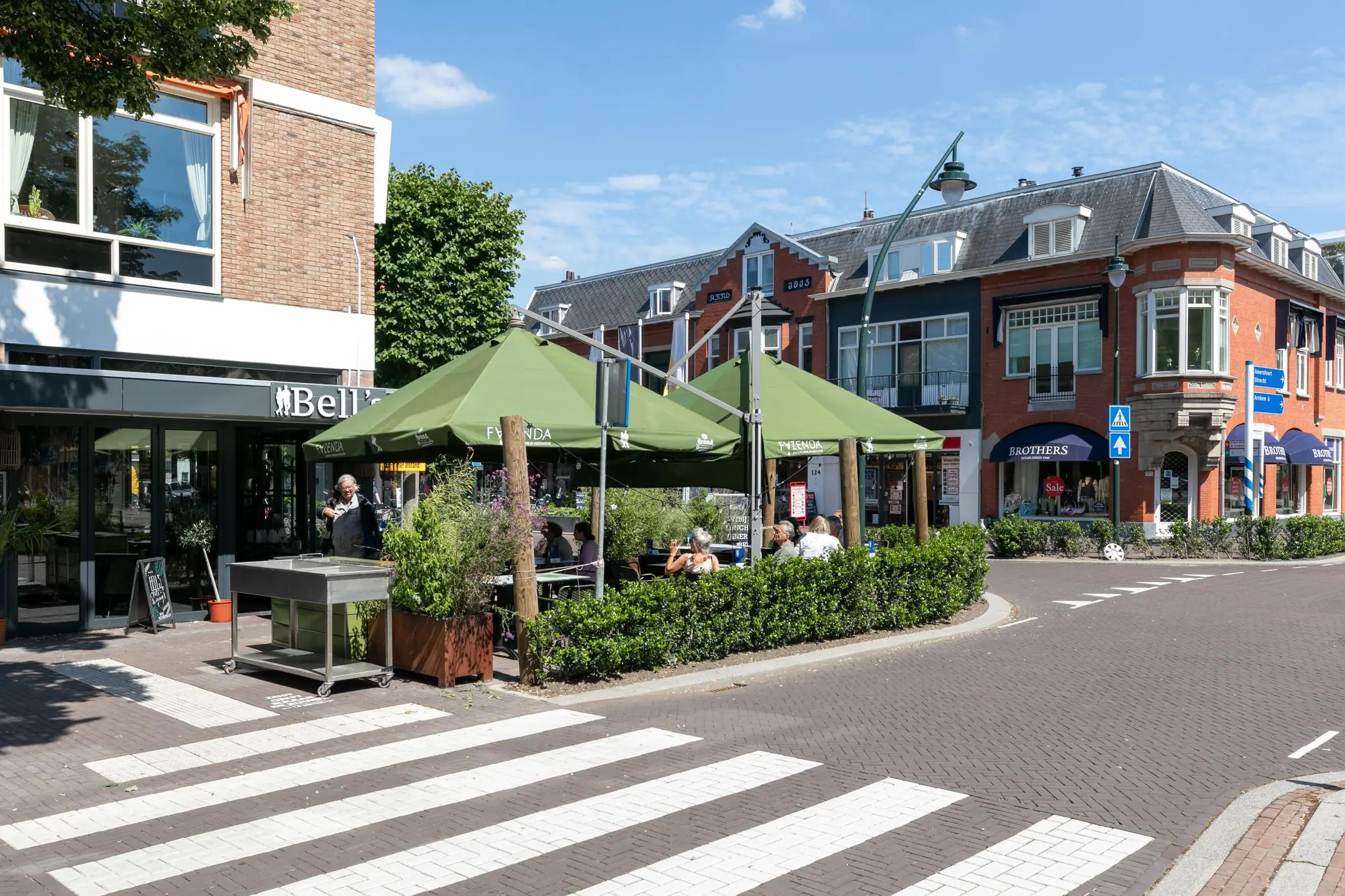 Terras met groene parasols aan de 1e Hogeweg in een levendige winkelstraat met restaurants en winkels.
